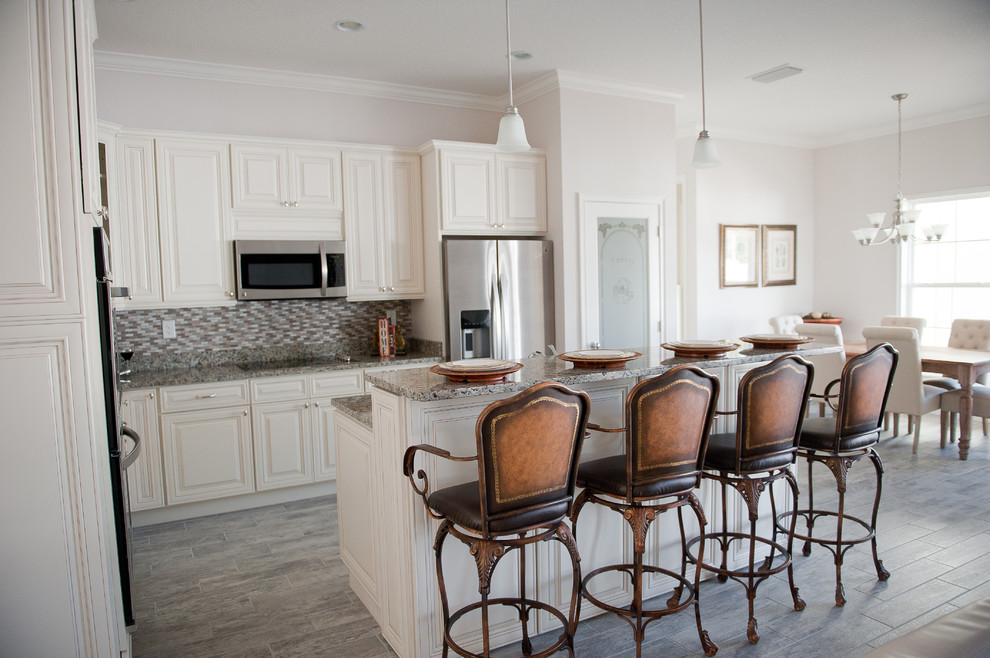 Mid-sized country l-shaped eat-in kitchen in Orlando with an undermount sink, raised-panel cabinets, granite benchtops, stainless steel appliances, porcelain floors, with island, white cabinets, multi-coloured splashback and matchstick tile splashback.