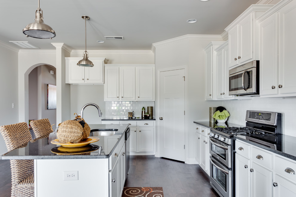 Mid-sized transitional l-shaped dark wood floor open concept kitchen photo with an undermount sink, recessed-panel cabinets, white cabinets, solid surface countertops, white backsplash, subway tile backsplash, stainless steel appliances and an island