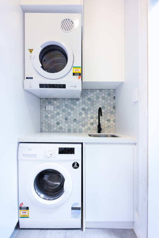 Photo of a small modern l-shaped utility room in Sydney with an undermount sink, flat-panel cabinets, white cabinets, quartz benchtops, multi-coloured walls, ceramic floors, a stacked washer and dryer, grey floor and white benchtop.