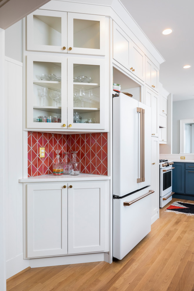 This is an example of a mid-sized eclectic l-shaped eat-in kitchen in Seattle with flat-panel cabinets, white cabinets, quartz benchtops, red splashback, ceramic splashback, white appliances, medium hardwood floors and white benchtop.