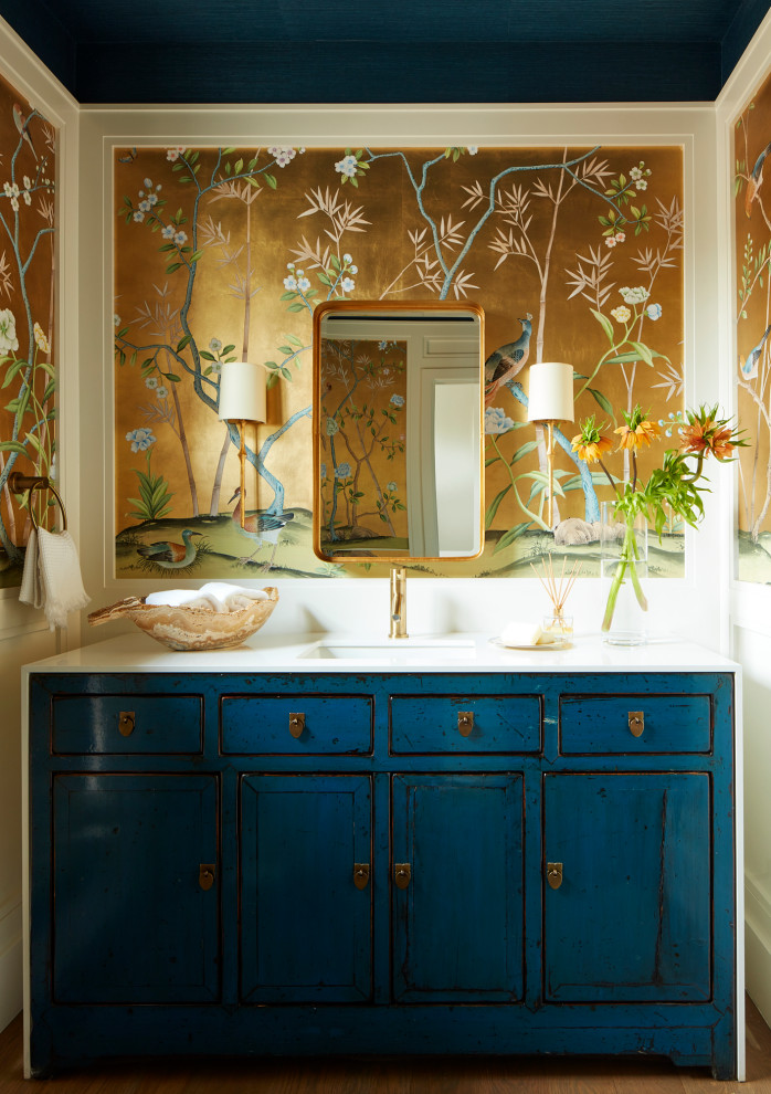 Modelo de aseo tradicional renovado con puertas de armario azules, suelo de madera clara, lavabo bajoencimera, encimera de mármol, suelo marrón, encimeras blancas, papel pintado y papel pintado