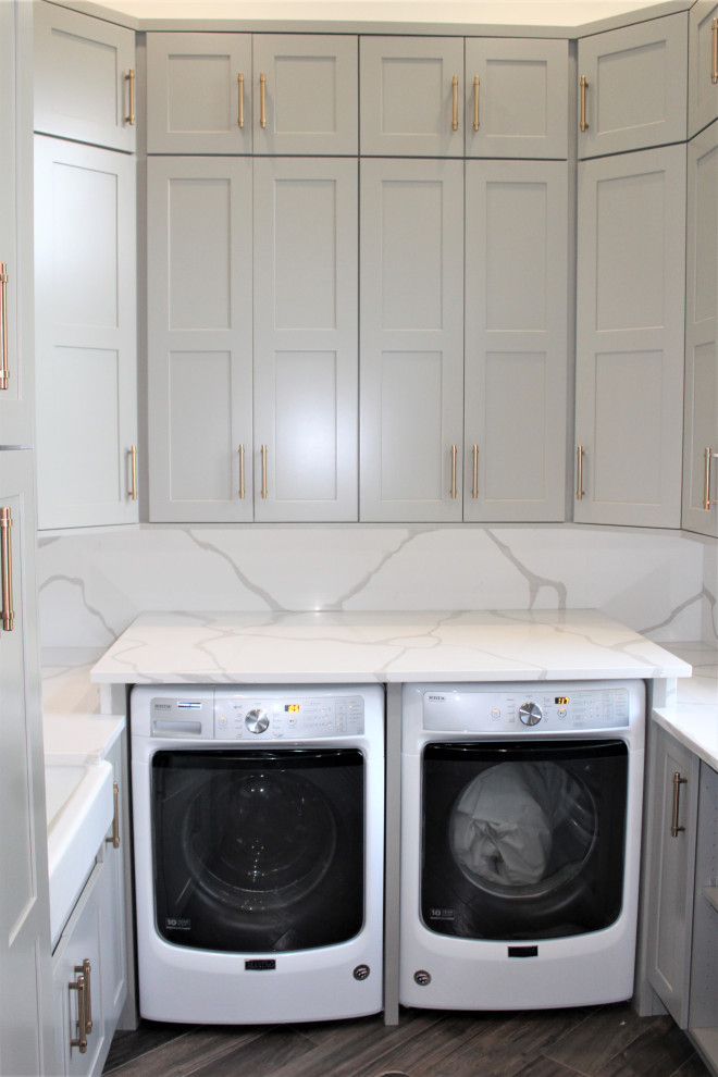 Inspiration for a large transitional u-shaped dedicated laundry room in Detroit with a farmhouse sink, shaker cabinets, grey cabinets, quartz benchtops, white splashback, engineered quartz splashback, grey walls, dark hardwood floors, a side-by-side washer and dryer, brown floor and white benchtop.