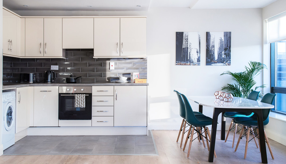 This is an example of a mid-sized contemporary l-shaped eat-in kitchen in Dublin with flat-panel cabinets, white cabinets, grey splashback, subway tile splashback, stainless steel appliances, light hardwood floors and grey floor.