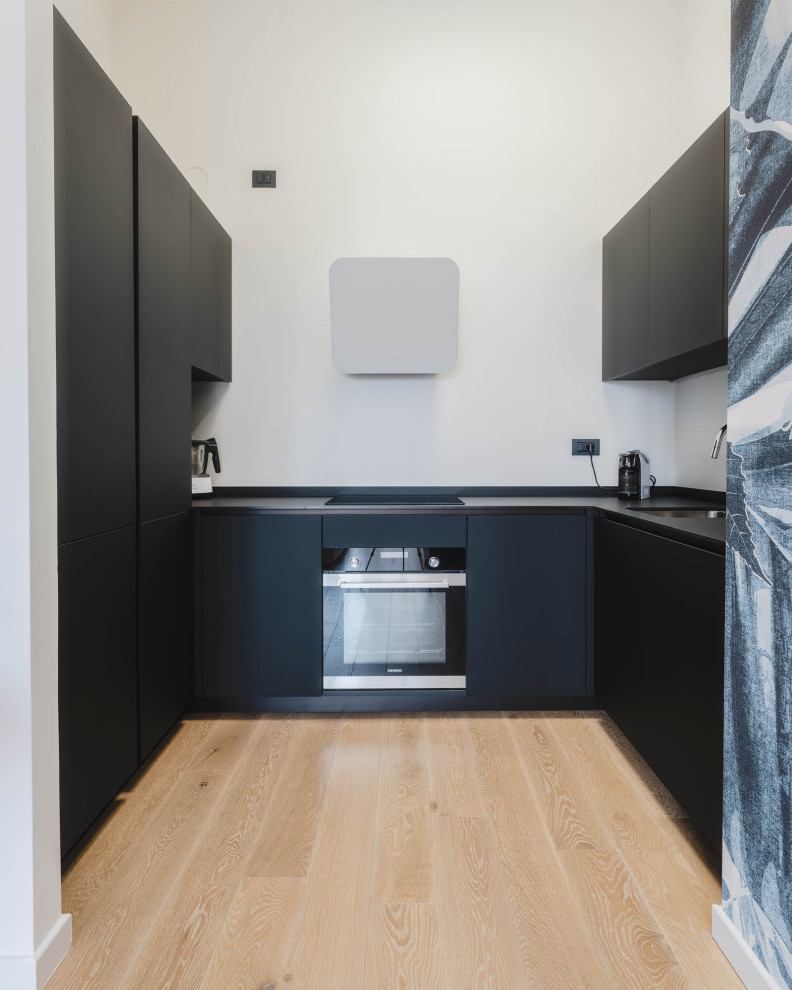 Small contemporary u-shaped kitchen/diner in Milan with a submerged sink, flat-panel cabinets, black cabinets, composite countertops, white splashback, integrated appliances, light hardwood flooring, no island, beige floors and black worktops.