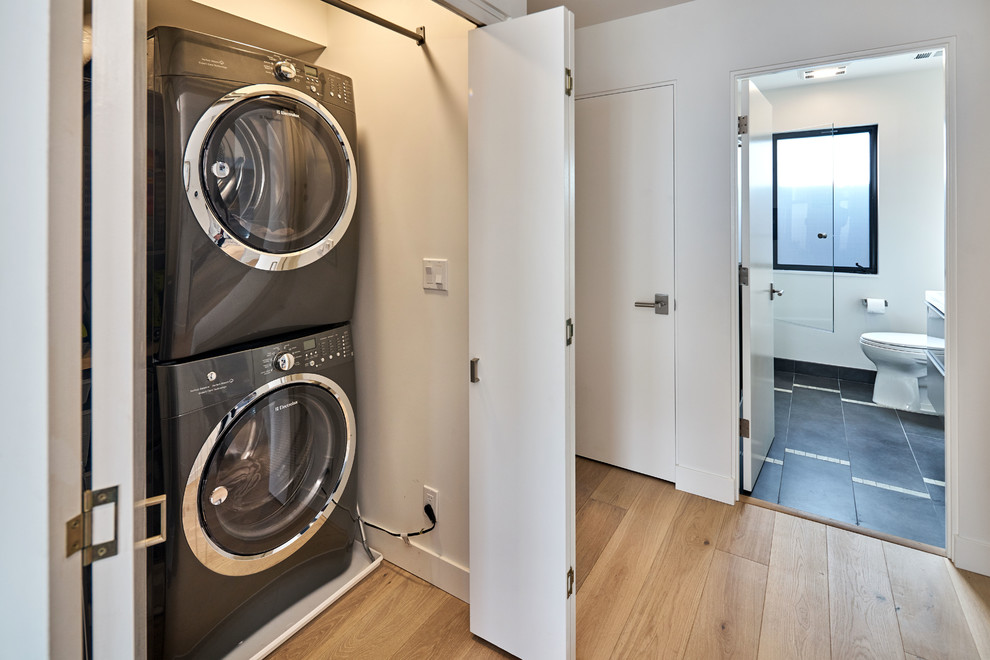 Design ideas for a small contemporary single-wall laundry cupboard in San Francisco with white walls, light hardwood floors and a stacked washer and dryer.