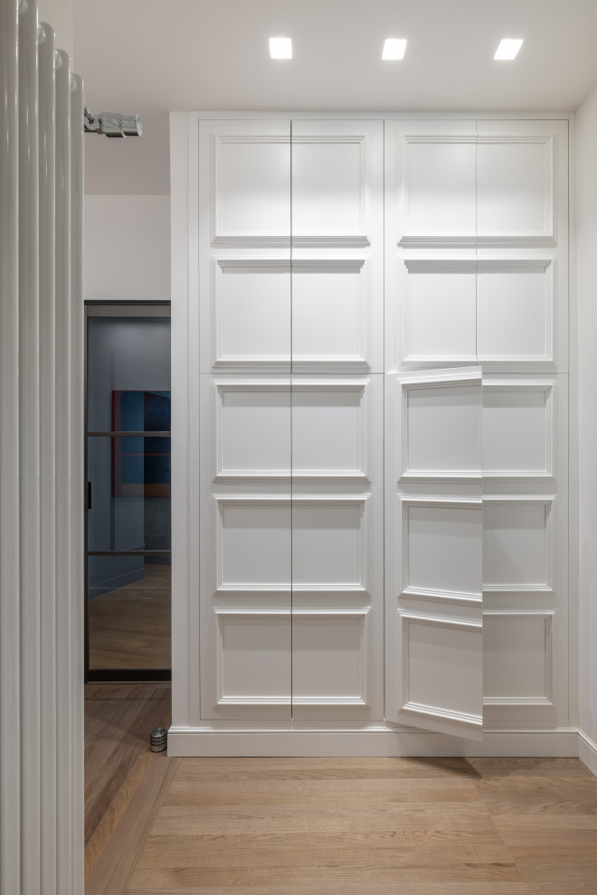 Cette photo montre un placard dressing moderne neutre avec un placard avec porte à panneau encastré, des portes de placard blanches, parquet clair et un sol marron.