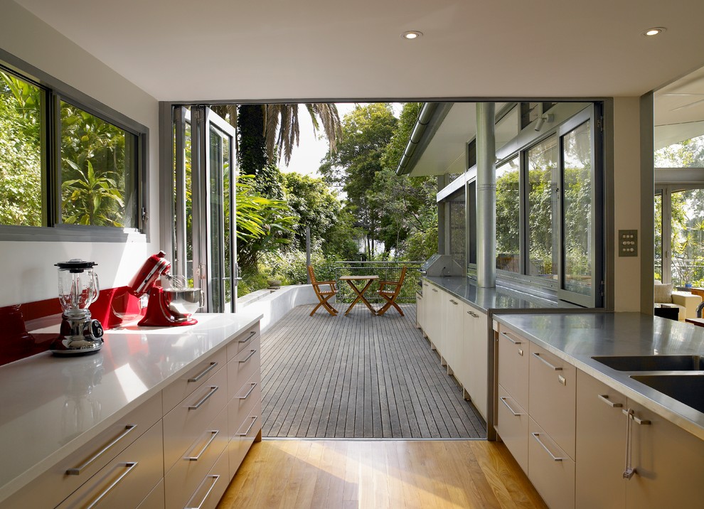 This is an example of a mid-sized contemporary galley eat-in kitchen in Sydney with an integrated sink, flat-panel cabinets, beige cabinets, stainless steel benchtops, red splashback, light hardwood floors, stainless steel appliances and a peninsula.