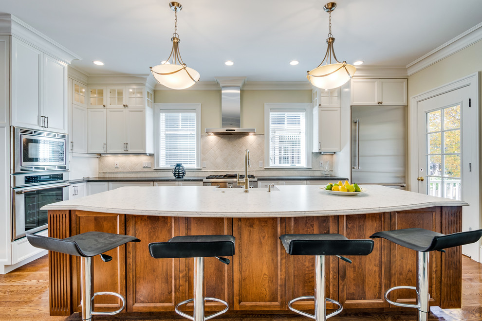 Example of a mid-sized transitional l-shaped medium tone wood floor and brown floor open concept kitchen design in Boston with a farmhouse sink, recessed-panel cabinets, white cabinets, soapstone countertops, beige backsplash, ceramic backsplash, stainless steel appliances and an island