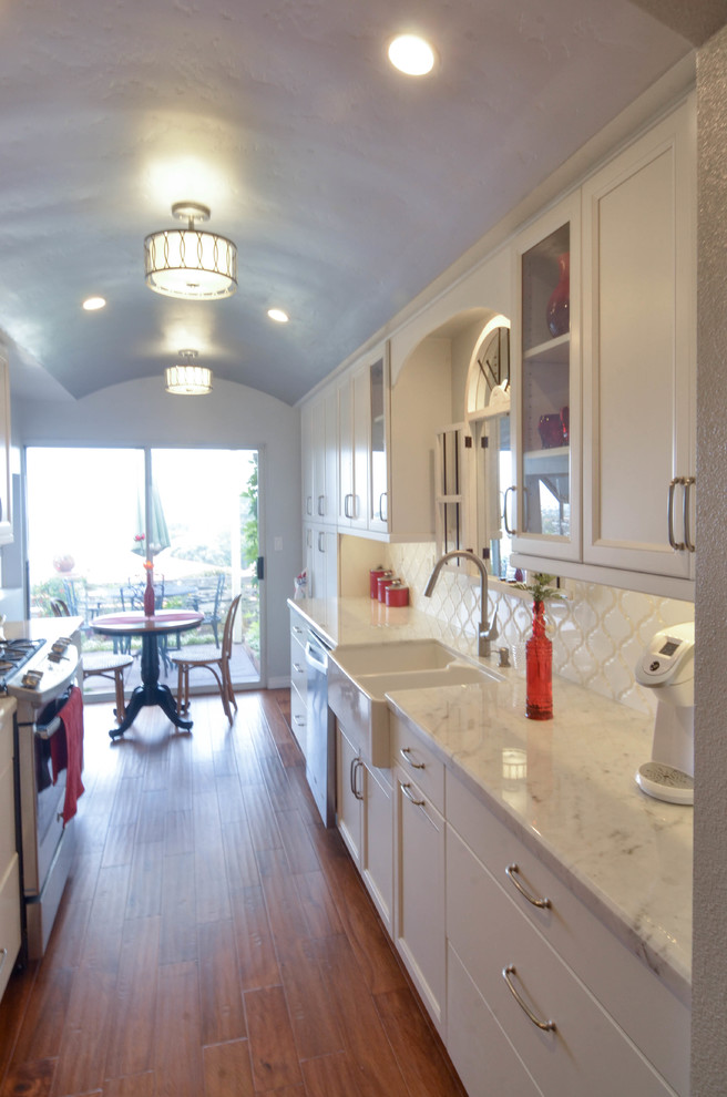 Photo of a mid-sized transitional galley eat-in kitchen in San Diego with a farmhouse sink, recessed-panel cabinets, white cabinets, marble benchtops, white splashback, porcelain splashback, stainless steel appliances, medium hardwood floors and no island.