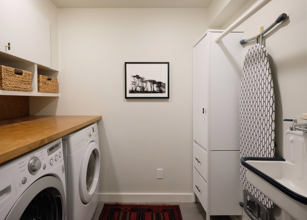 This is an example of a mid-sized contemporary galley dedicated laundry room in San Francisco with a farmhouse sink, flat-panel cabinets, white cabinets, wood benchtops, white walls, concrete floors, a side-by-side washer and dryer and grey floor.