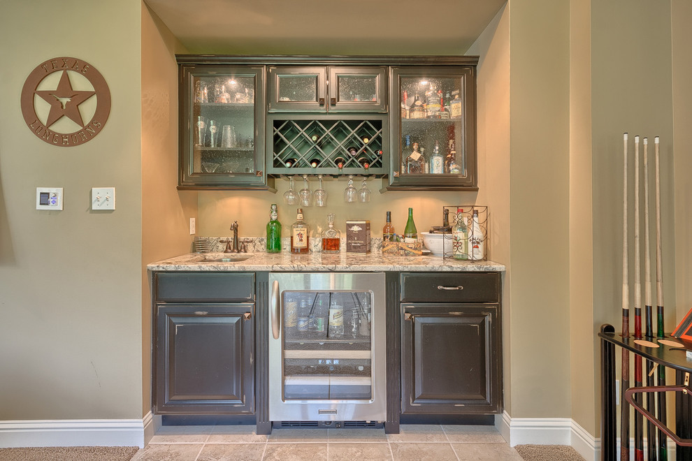 Photo of a small traditional single-wall wet bar in St Louis with an undermount sink, dark wood cabinets, granite benchtops, ceramic floors, raised-panel cabinets and beige floor.