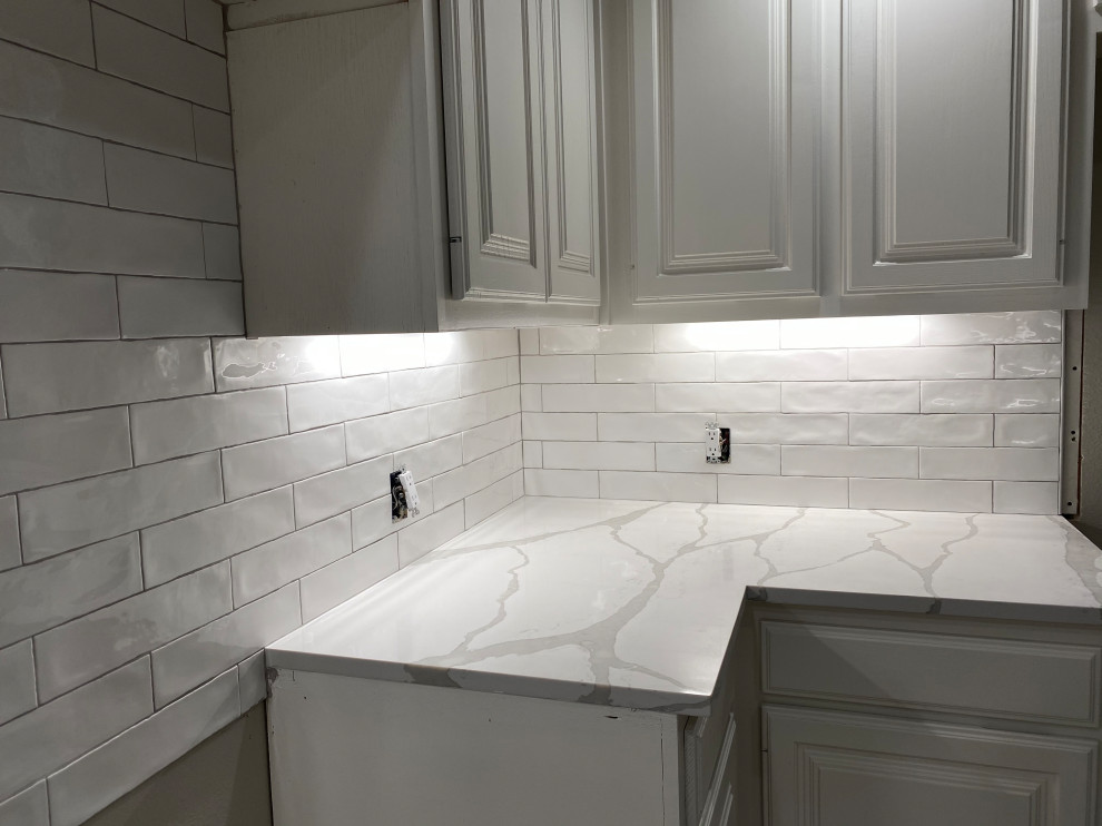 Photo of a mid-sized transitional l-shaped eat-in kitchen in Austin with a single-bowl sink, raised-panel cabinets, white cabinets, quartz benchtops, white splashback, ceramic splashback, white appliances and white benchtop.