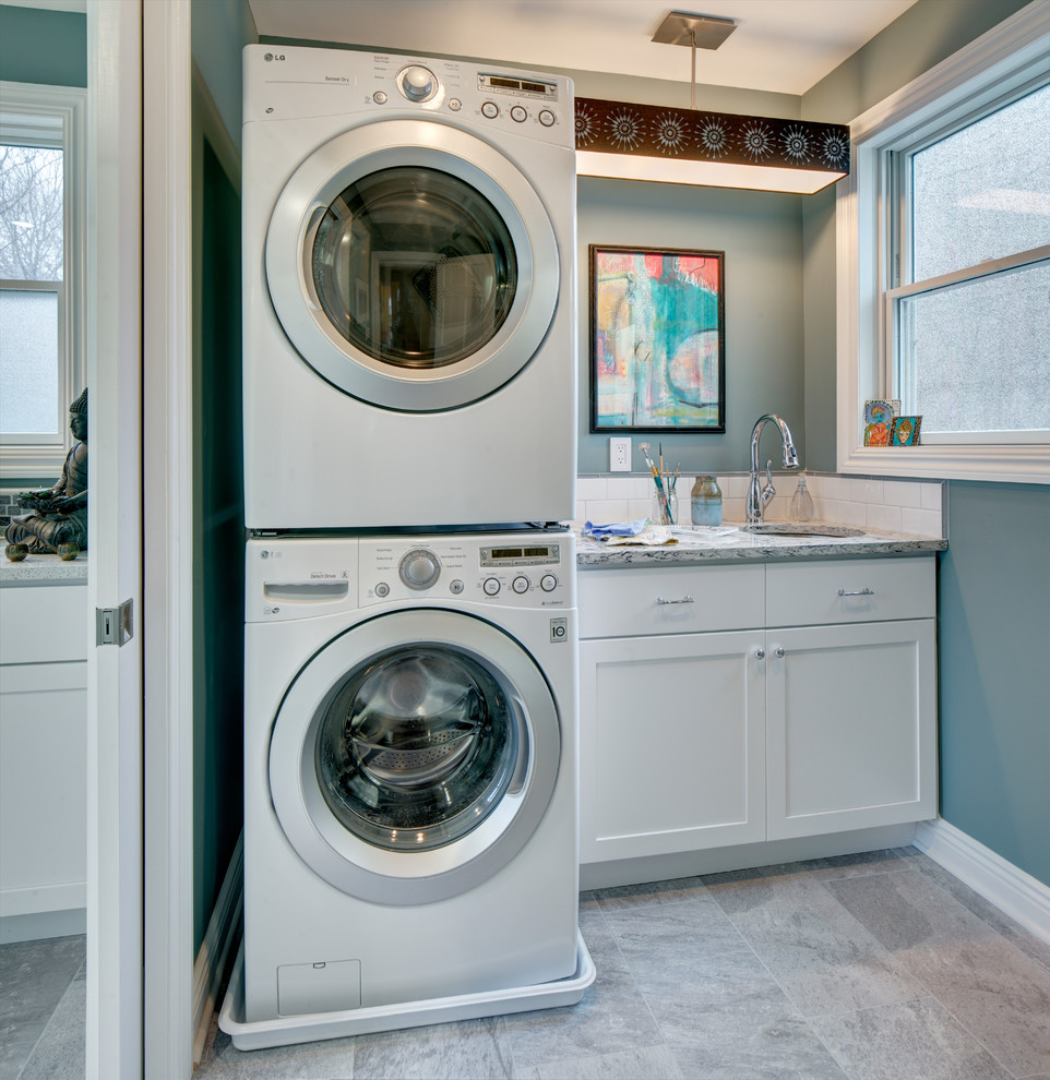 This is an example of a small transitional single-wall dedicated laundry room in New York with an undermount sink, shaker cabinets, white cabinets, quartz benchtops, porcelain floors, a stacked washer and dryer, grey floor and blue walls.