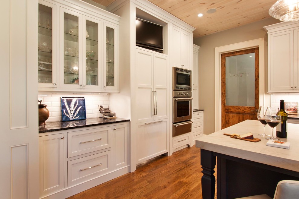 Inspiration for a mid-sized transitional l-shaped light wood floor and brown floor eat-in kitchen remodel with glass-front cabinets, white cabinets, granite countertops, white backsplash, ceramic backsplash, stainless steel appliances, an island and multicolored countertops