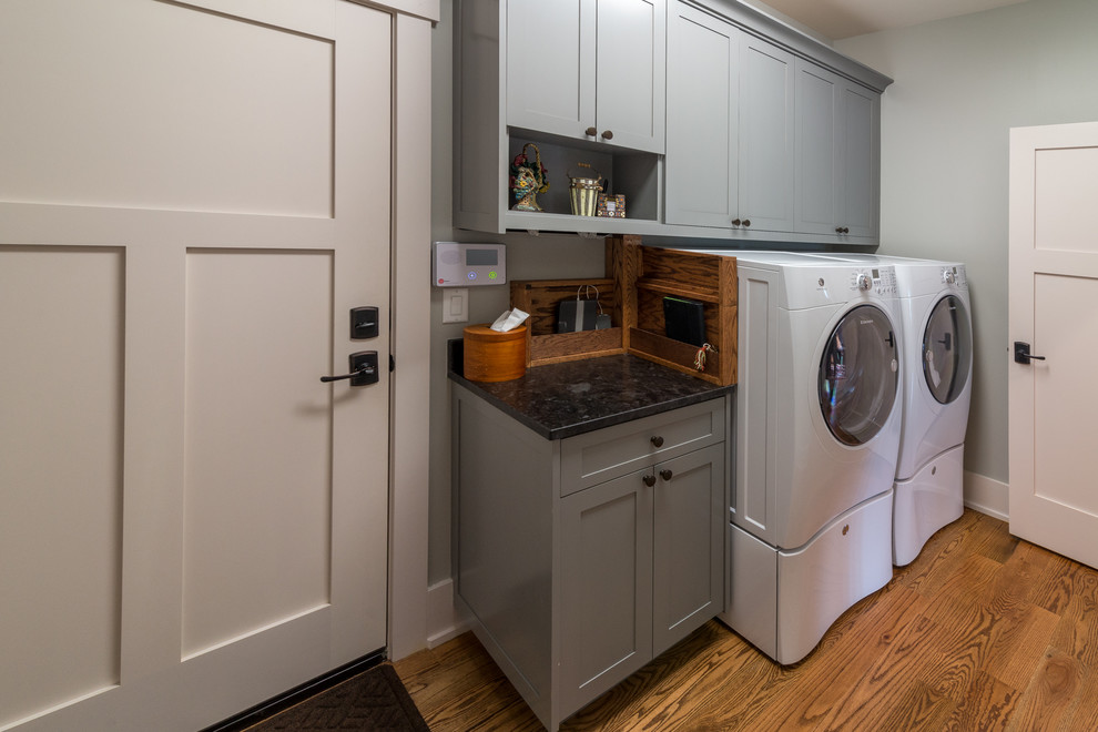 Photo of a mid-sized transitional single-wall dedicated laundry room in Other with shaker cabinets, grey cabinets, soapstone benchtops, grey walls, medium hardwood floors, a side-by-side washer and dryer, brown floor and black benchtop.