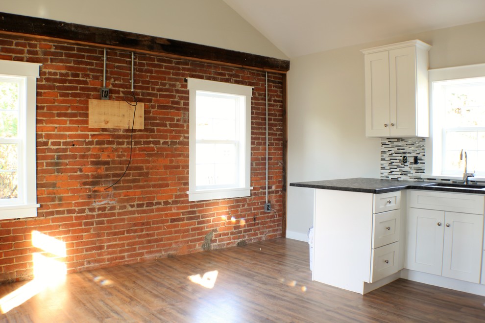 Example of a small trendy single-wall medium tone wood floor open concept kitchen design with an undermount sink, recessed-panel cabinets, white cabinets, granite countertops, gray backsplash, glass tile backsplash, stainless steel appliances and a peninsula