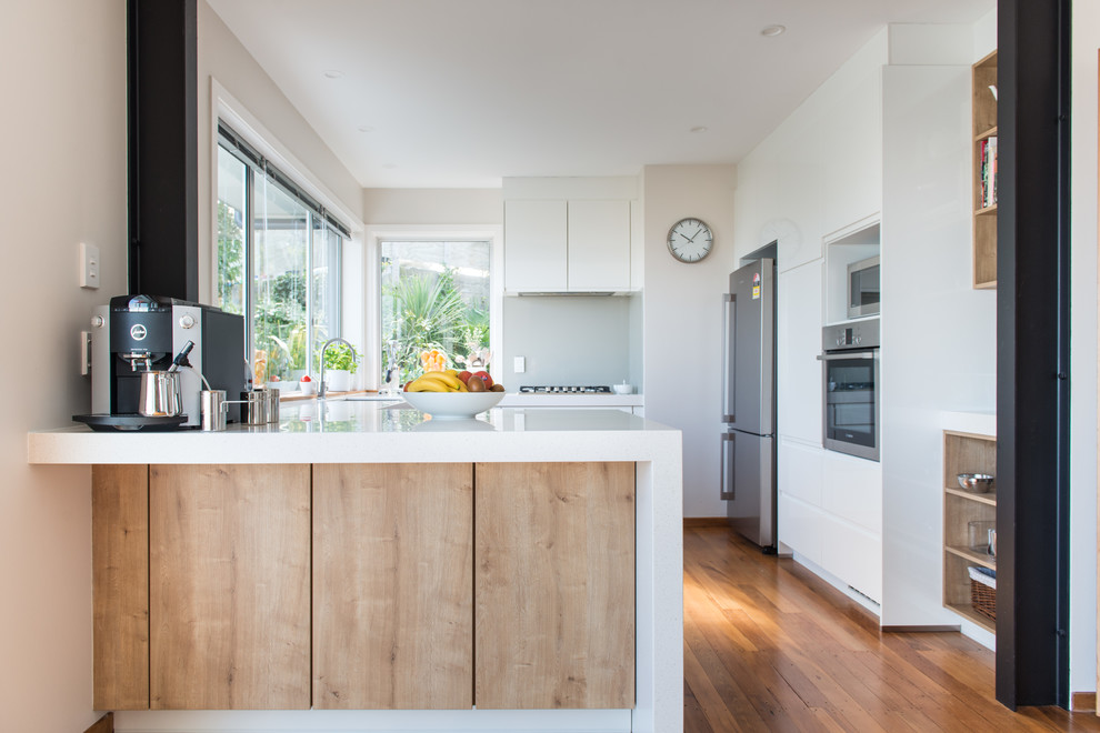 Photo of a mid-sized scandinavian u-shaped kitchen pantry in Christchurch with an undermount sink, flat-panel cabinets, white cabinets, solid surface benchtops, glass sheet splashback, white appliances, medium hardwood floors, a peninsula and brown floor.