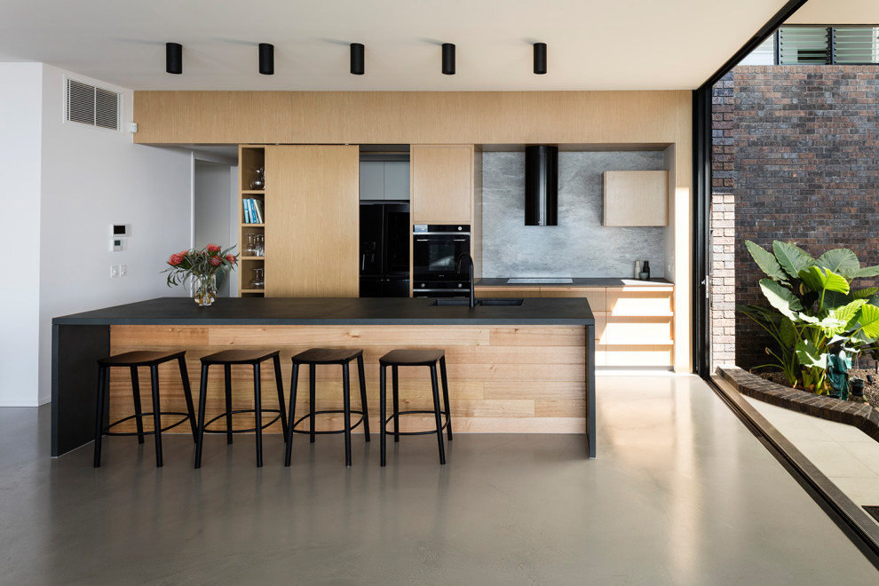 Inspiration for a modern galley kitchen in Sunshine Coast with an undermount sink, flat-panel cabinets, light wood cabinets, grey splashback, stone slab splashback, black appliances, concrete floors, with island, grey floor and black benchtop.