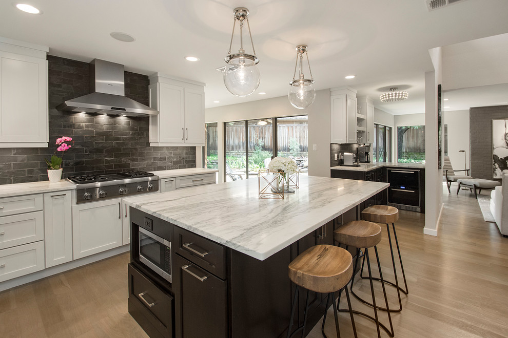 This is an example of a large modern u-shaped open plan kitchen in Dallas with a farmhouse sink, shaker cabinets, white cabinets, marble benchtops, black splashback, stone tile splashback, stainless steel appliances, light hardwood floors and with island.