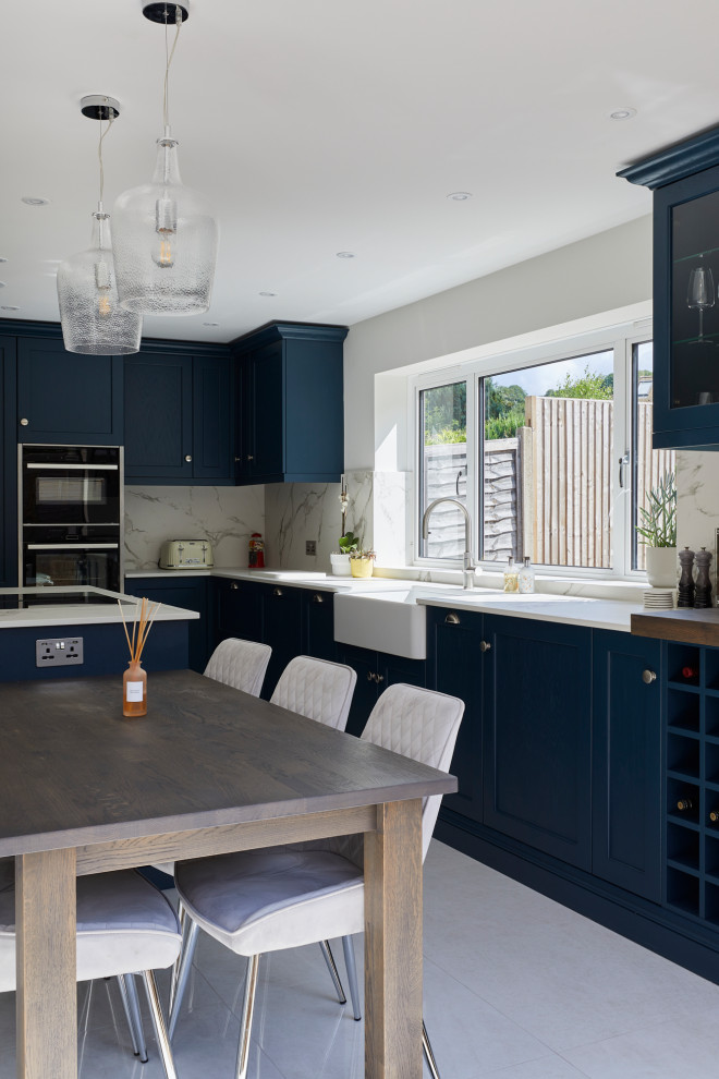 Photo of a large contemporary l-shaped eat-in kitchen in London with a farmhouse sink, recessed-panel cabinets, blue cabinets, quartz benchtops, white splashback, stone slab splashback, stainless steel appliances, with island and white benchtop.