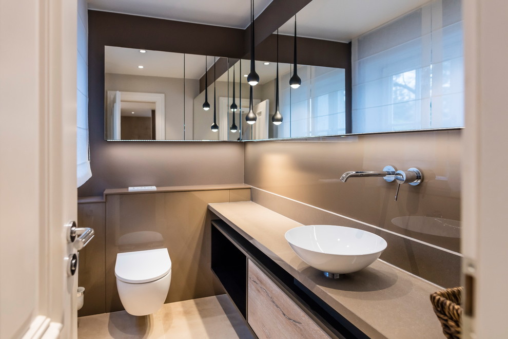 This is an example of a contemporary powder room in Frankfurt with flat-panel cabinets, light wood cabinets, a wall-mount toilet, beige tile, brown walls, a vessel sink, beige floor and beige benchtops.