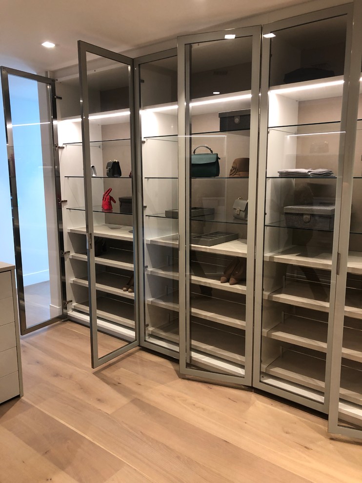 Photo of an expansive modern gender-neutral walk-in wardrobe in Miami with glass-front cabinets, white cabinets, light hardwood floors and beige floor.