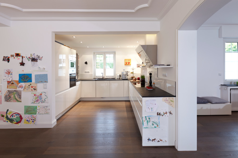 This is an example of a mid-sized contemporary u-shaped open plan kitchen in Essen with an integrated sink, flat-panel cabinets, white cabinets, white splashback, glass sheet splashback, dark hardwood floors and no island.
