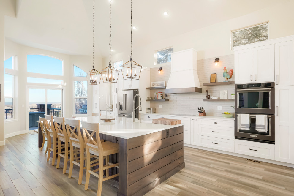 This is an example of a large transitional single-wall open plan kitchen in Denver with a farmhouse sink, shaker cabinets, white cabinets, white splashback, subway tile splashback, black appliances, with island, white benchtop and vaulted.