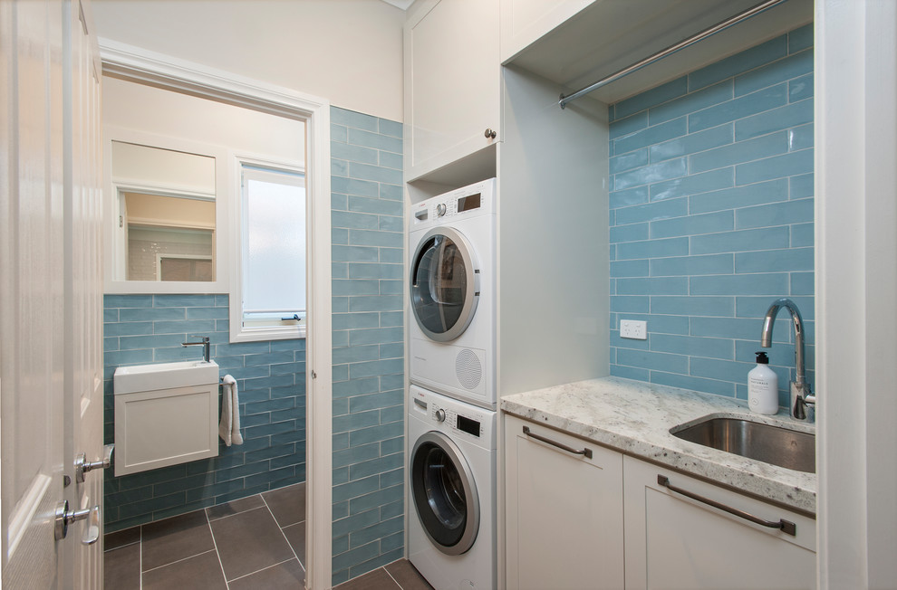 This is an example of a small transitional single-wall utility room in Melbourne with an undermount sink, shaker cabinets, beige cabinets, granite benchtops, porcelain floors, a stacked washer and dryer, grey floor, beige walls, beige benchtop, ceramic splashback and blue splashback.