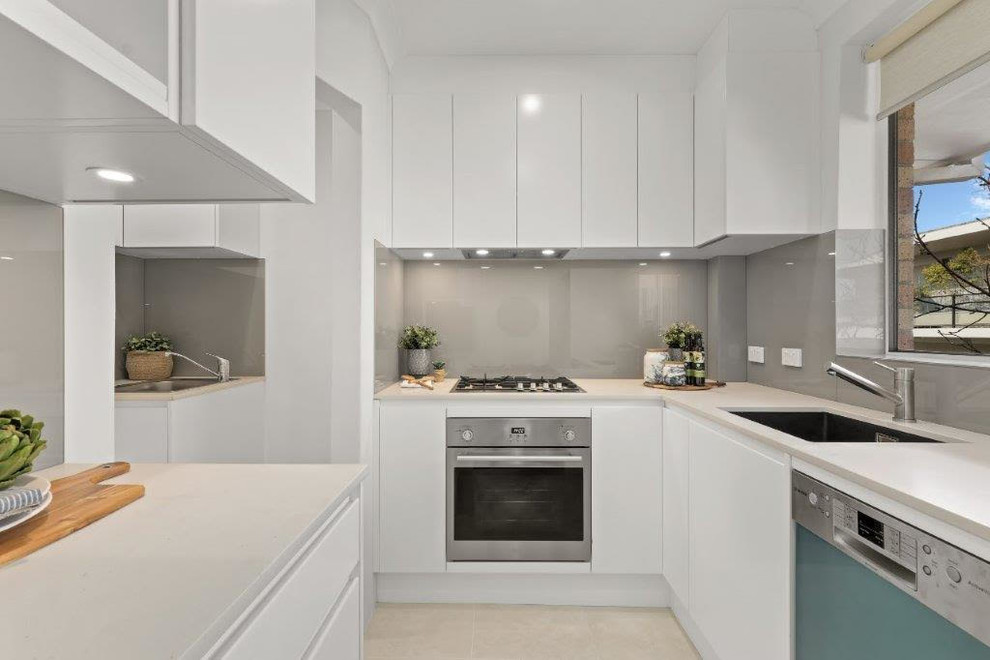 Photo of a small beach style u-shaped separate kitchen in Sydney with an undermount sink, flat-panel cabinets, white cabinets, quartz benchtops, grey splashback, glass sheet splashback, stainless steel appliances, ceramic floors, beige floor and white benchtop.