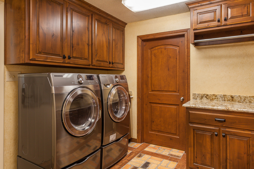 Mid-sized country u-shaped dedicated laundry room in Dallas with raised-panel cabinets, medium wood cabinets, granite benchtops, beige walls, brick floors and a side-by-side washer and dryer.