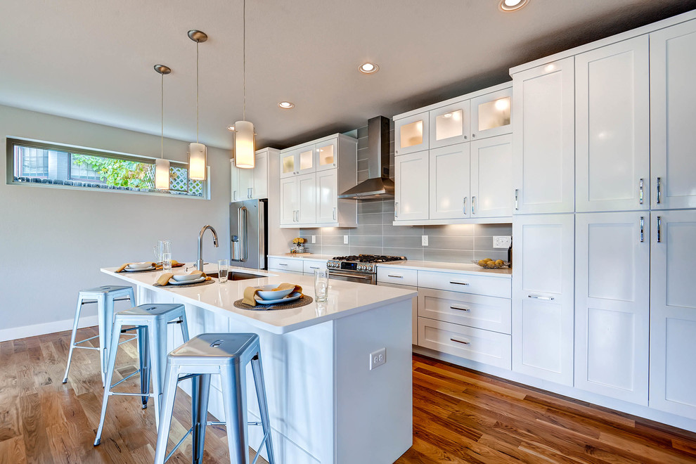 Design ideas for a transitional galley open plan kitchen in Denver with an undermount sink, shaker cabinets, white cabinets, grey splashback, stainless steel appliances, medium hardwood floors and with island.