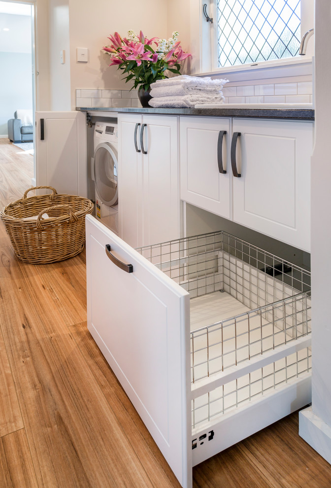 Photo of a mid-sized transitional u-shaped laundry room in Christchurch with white cabinets, granite benchtops, white walls, medium hardwood floors, a concealed washer and dryer, black benchtop, an undermount sink, shaker cabinets and beige floor.