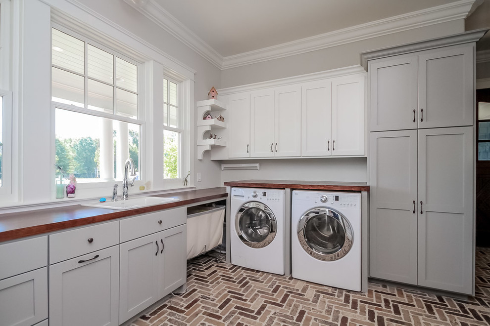 Photo of a large transitional l-shaped dedicated laundry room in Charlotte with a drop-in sink, shaker cabinets, grey cabinets, wood benchtops, grey walls, a side-by-side washer and dryer and brown floor.