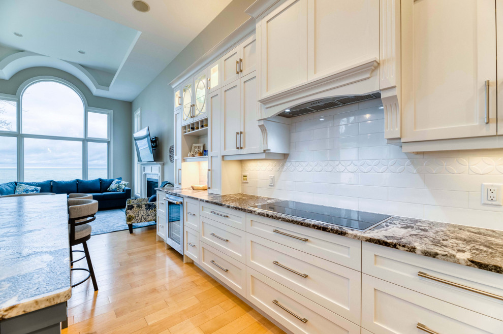 Large transitional l-shaped light wood floor and brown floor eat-in kitchen photo with a farmhouse sink, shaker cabinets, white cabinets, granite countertops, ceramic backsplash, stainless steel appliances and an island