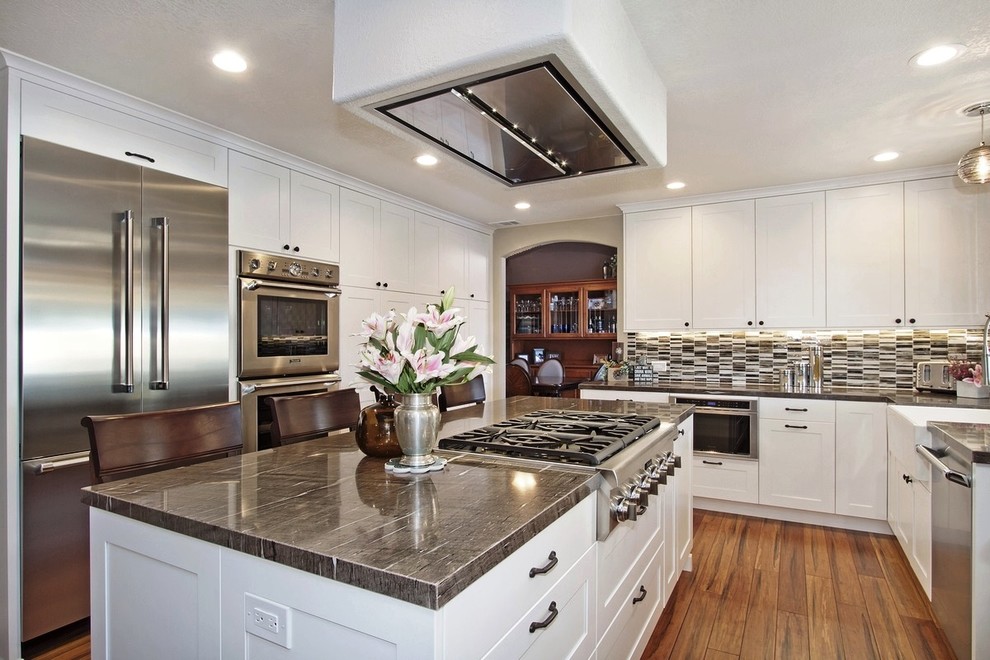 This is an example of a mid-sized transitional l-shaped eat-in kitchen in San Diego with a farmhouse sink, flat-panel cabinets, white cabinets, granite benchtops, multi-coloured splashback, glass tile splashback, stainless steel appliances, medium hardwood floors, with island and brown floor.