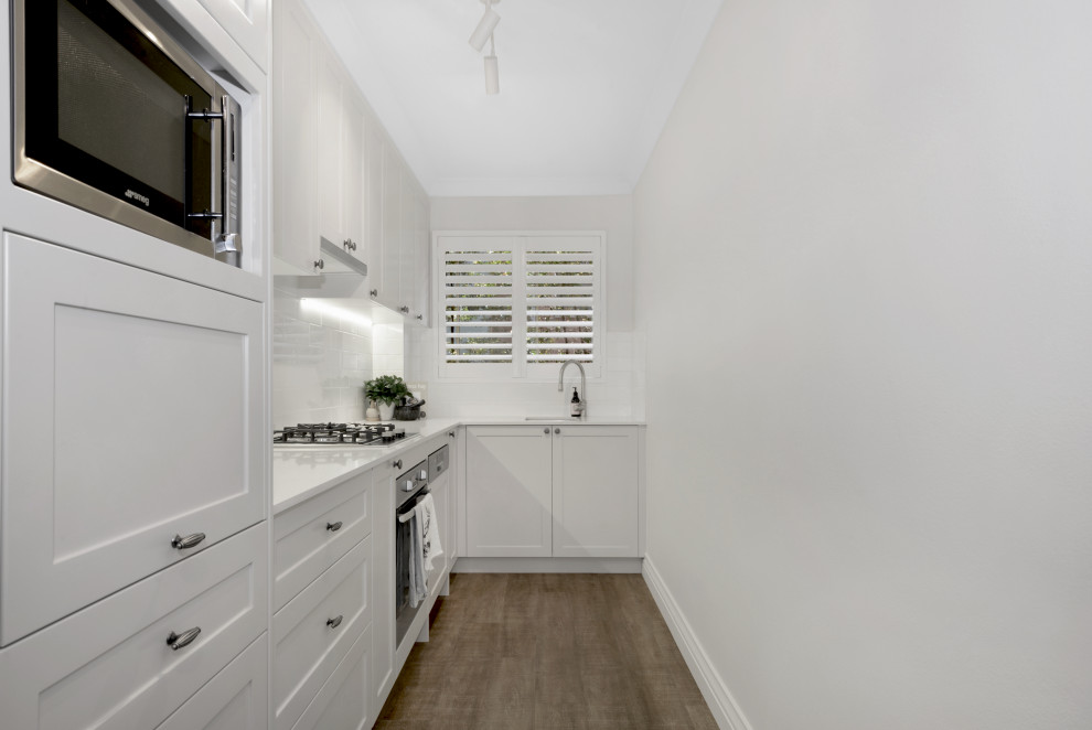 This is an example of a transitional l-shaped kitchen in Sydney with an undermount sink, shaker cabinets, white cabinets, white splashback, glass tile splashback, stainless steel appliances, light hardwood floors, no island, brown floor and white benchtop.