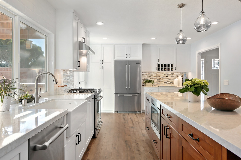Inspiration for a large transitional l-shaped kitchen in Tampa with a farmhouse sink, shaker cabinets, white cabinets, quartz benchtops, stainless steel appliances, vinyl floors, with island, brown floor, white benchtop, multi-coloured splashback and mosaic tile splashback.