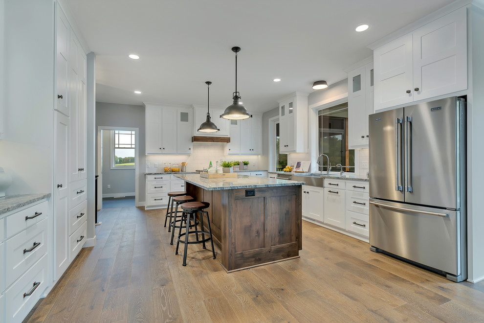 Photo of a mid-sized arts and crafts u-shaped eat-in kitchen in Minneapolis with a farmhouse sink, shaker cabinets, white cabinets, granite benchtops, white splashback, ceramic splashback, stainless steel appliances, medium hardwood floors, with island, brown floor and grey benchtop.
