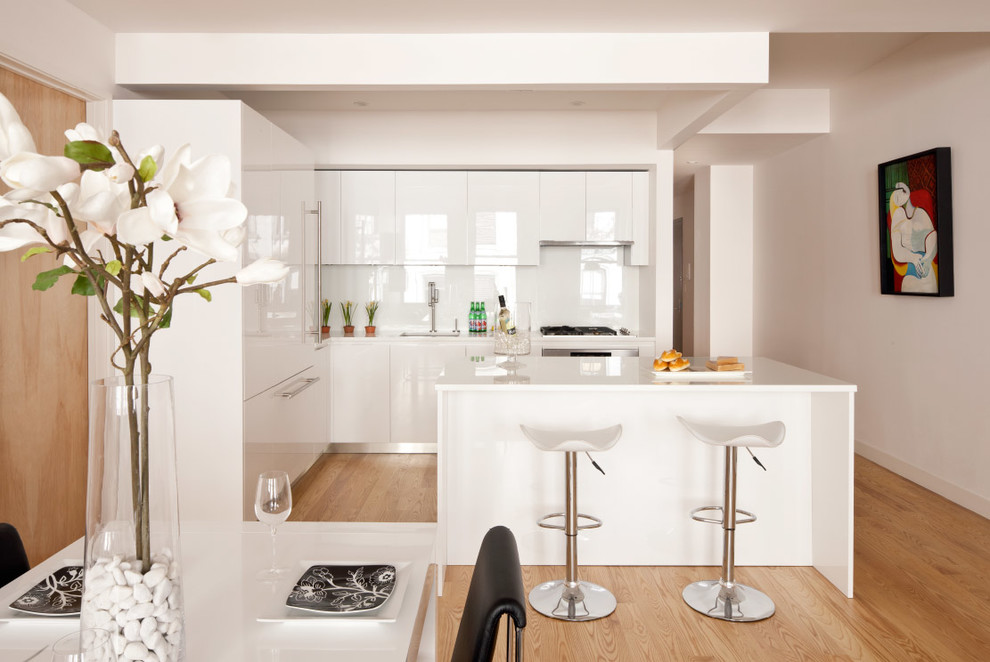 Contemporary l-shaped eat-in kitchen in New York with flat-panel cabinets, white cabinets, white splashback, glass sheet splashback and panelled appliances.