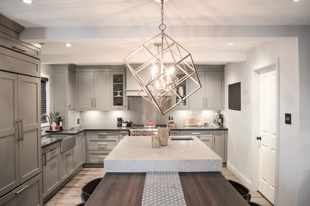 Mid-sized transitional l-shaped medium tone wood floor enclosed kitchen photo in Toronto with a farmhouse sink, shaker cabinets, gray cabinets, quartz countertops, gray backsplash, porcelain backsplash, paneled appliances and an island