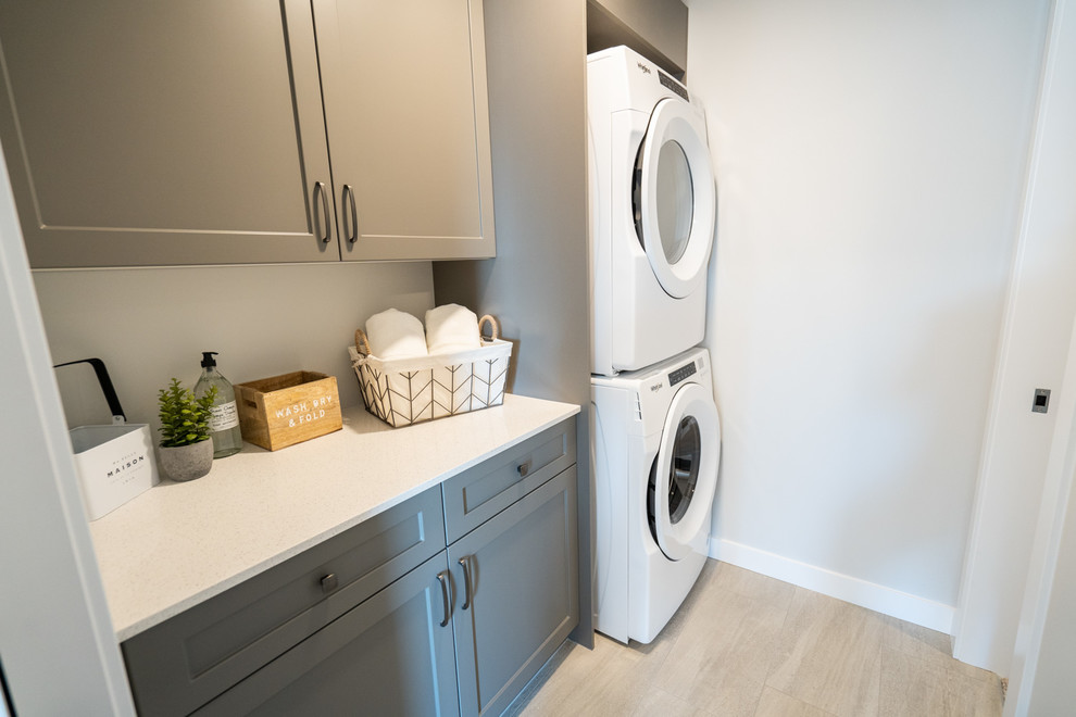 Inspiration for a mid-sized transitional single-wall dedicated laundry room in Edmonton with shaker cabinets, grey cabinets, quartz benchtops, grey walls, porcelain floors, a stacked washer and dryer, beige floor and white benchtop.