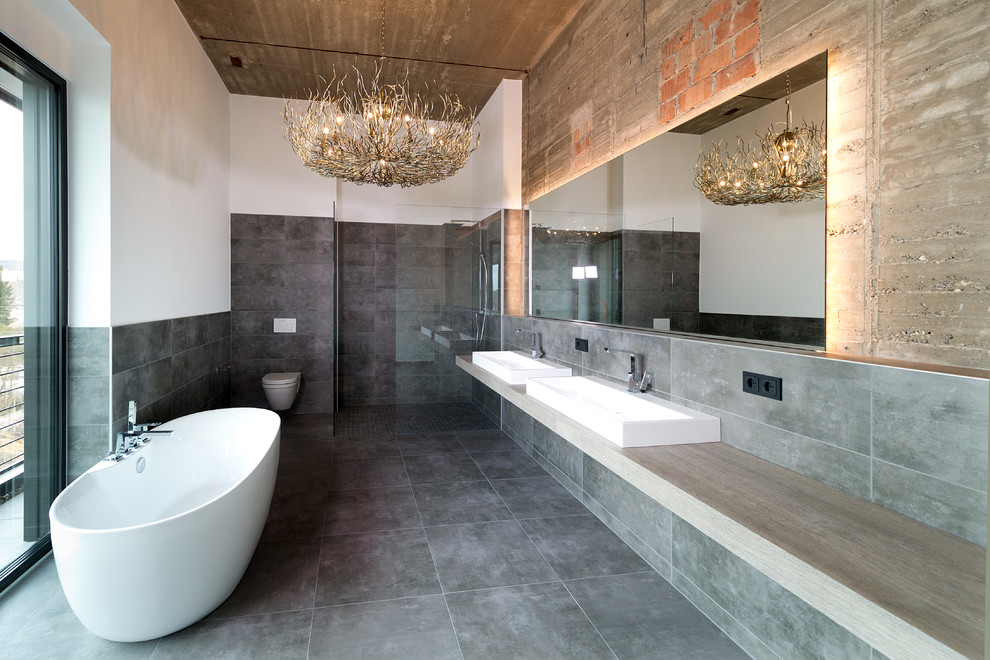 Großes Industrial Badezimmer En Suite mit freistehender Badewanne, Keramikboden, Aufsatzwaschbecken, Eckdusche, Wandtoilette, grauer Wandfarbe, grauem Boden, offener Dusche, grauen Fliesen, Waschtisch aus Holz und grauer Waschtischplatte in Sonstige