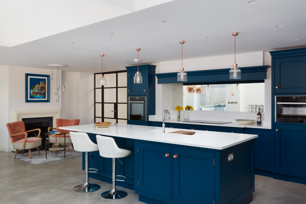 This is an example of a transitional u-shaped open plan kitchen in London with an undermount sink, shaker cabinets, blue cabinets, white splashback, mirror splashback, black appliances, concrete floors, with island, grey floor and white benchtop.