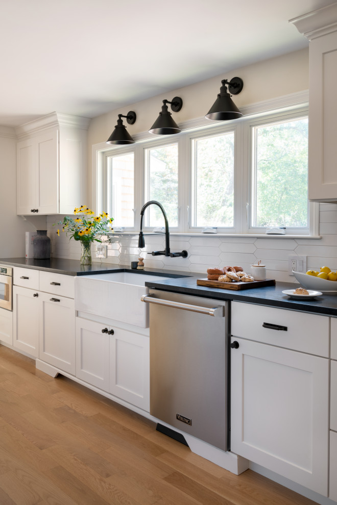 Mid-sized country galley eat-in kitchen in Boston with a farmhouse sink, shaker cabinets, white cabinets, granite benchtops, white splashback, porcelain splashback, stainless steel appliances, light hardwood floors, no island, beige floor and black benchtop.