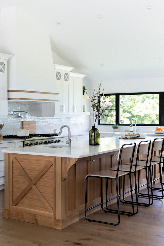 Photo of an expansive beach style l-shaped eat-in kitchen in Toronto with an undermount sink, shaker cabinets, white cabinets, quartz benchtops, white splashback, subway tile splashback, stainless steel appliances, light hardwood floors, with island, brown floor and white benchtop.
