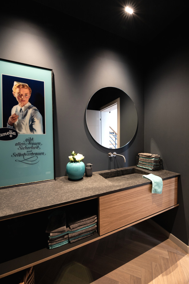 Inspiration for a mid-sized contemporary powder room in Other with flat-panel cabinets, black walls, light hardwood floors, an integrated sink, granite benchtops, beige floor, black benchtops, medium wood cabinets, a two-piece toilet and a floating vanity.