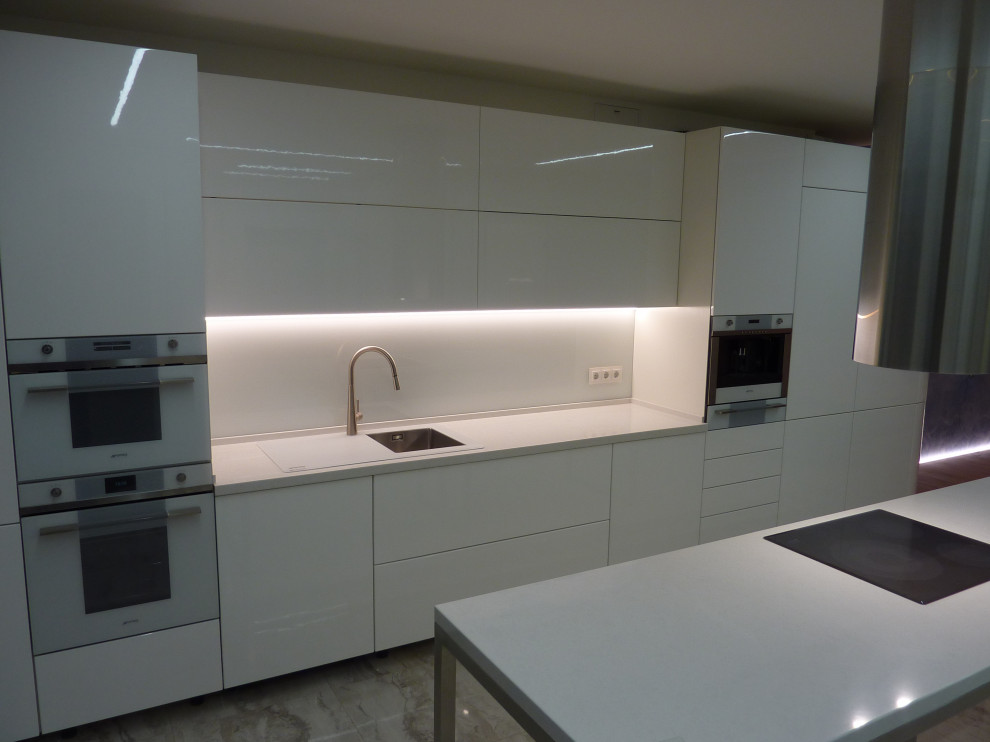 Inredning av ett modernt stort vit linjärt vitt kök och matrum, med en nedsänkt diskho, släta luckor, vita skåp, bänkskiva i kvarts, vitt stänkskydd, glaspanel som stänkskydd, vita vitvaror, en köksö och beiget golv