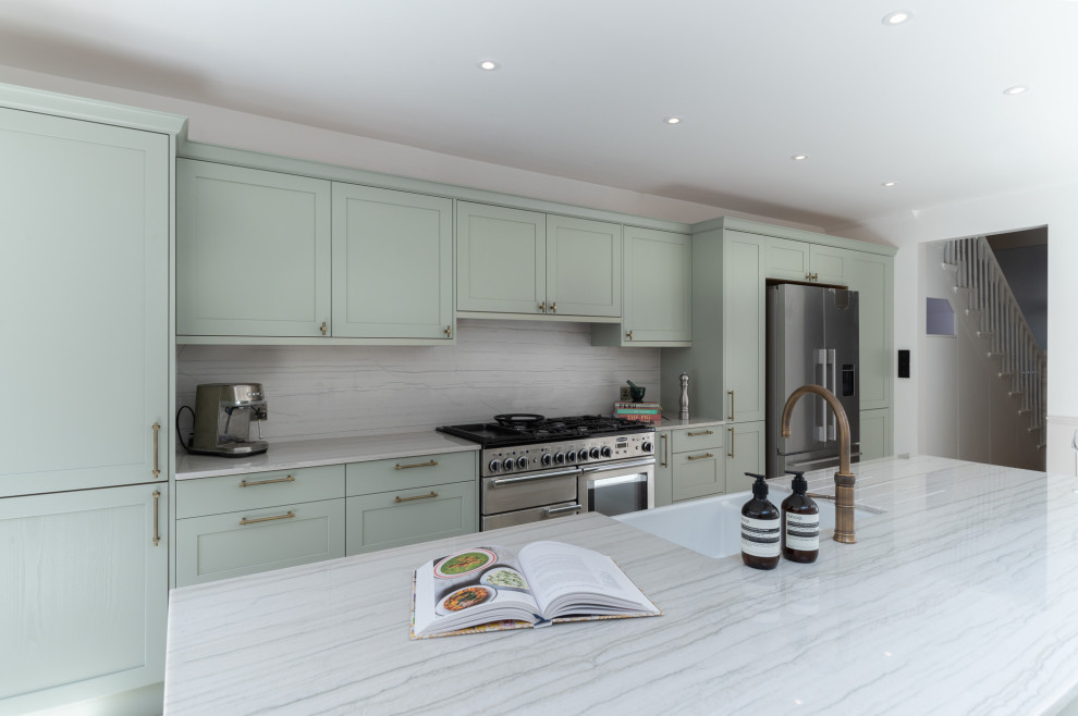 Photo of a large single-wall open plan kitchen in London with an integrated sink, shaker cabinets, green cabinets, marble benchtops, medium hardwood floors, with island, white benchtop, white splashback, engineered quartz splashback and stainless steel appliances.