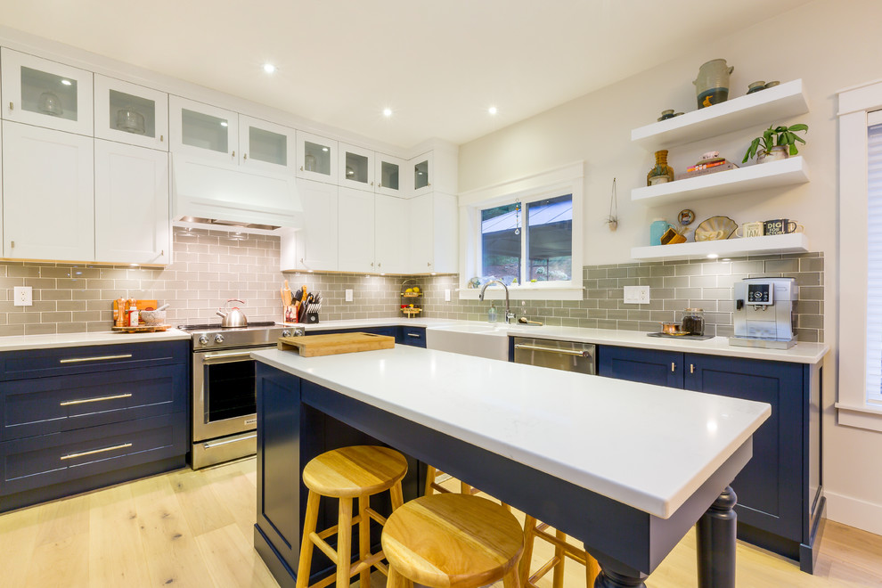 Large transitional u-shaped light wood floor open concept kitchen photo with a farmhouse sink, shaker cabinets, blue cabinets, quartz countertops, gray backsplash, glass tile backsplash, stainless steel appliances, an island and white countertops